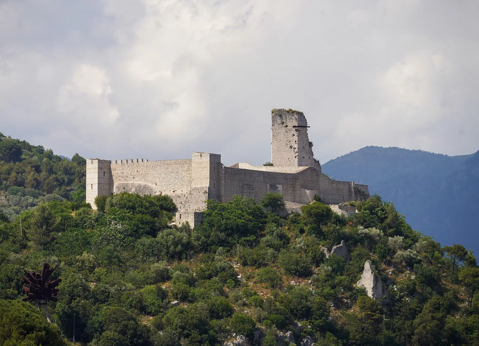 Rocca Janula, fortezza medievale in pietra chiara con mura merlate e una torre, sul fianco boscoso di Montecassino sotto un cielo nuvoloso
